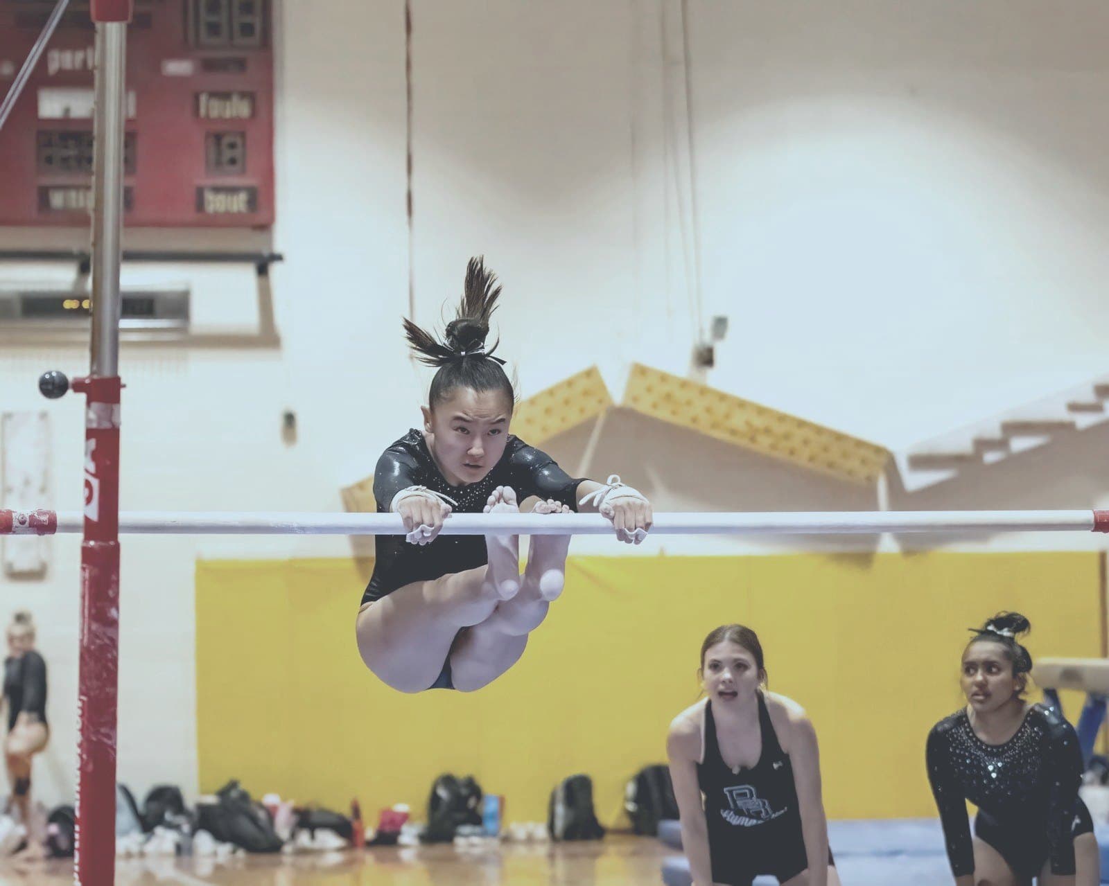 Gymnast grips the bar with complete focus, teammates watching